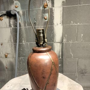 May include: A brown ceramic table lamp with a gold-colored metal accent design. The lamp has a brass-colored neck and a black power cord. The lamp sits on a gray surface against a gray brick wall.