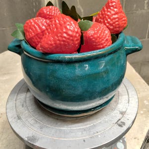 May include: A deep teal ceramic bowl with two side handles, filled with several bright red artificial strawberries featuring green fabric leaves. The bowl has a smooth, glossy finish and sits on a matching circular ceramic saucer base.