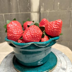 May include: A teal ceramic bowl with a scalloped rim and matching saucer, filled with several bright red, textured artificial strawberries. Each strawberry features small green paper leaves at the top, contrasting against the deep blue-green glaze.