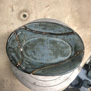 May include: An oval ceramic dish with a blue-grey speckled glaze, featuring prominent gold-colored lines that appear to repair cracks across the surface. The bowl has a smooth, glossy finish and a slightly irregular, organic shape.