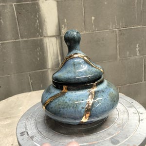 Pode incluir: Um pote de cerâmica azul com tampa correspondente, apresentando um acabamento brilhante e detalhes decorativos de cor dourada que traçam linhas irregulares na superfície da tampa e do corpo arredondado do recipiente.