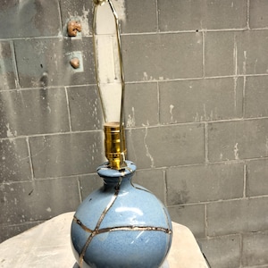 May include: A blue ceramic lamp base with gold accents, featuring a Kintsugi design. The lamp has a gold-colored neck and a tall, rectangular shade. The lamp is on a gray surface.