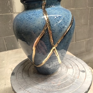 May include: A blue ceramic vase with a speckled finish and a glossy glaze. The vase features prominent gold-colored lines that traverse the surface in a pattern reminiscent of kintsugi, highlighting repaired or decorative cracks across the body.