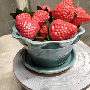 May include: A teal ceramic bowl with a scalloped rim and matching saucer, filled with multiple glossy red artificial strawberries featuring green fabric leaves. The bowl has a smooth, glazed finish and sits on a circular metal pottery wheel.