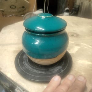 May include: A teal ceramic jar with a matching lid, sitting on a black circular base. The jar has a rounded shape with a lighter, natural clay base. The lid sits snugly on top, creating a cohesive design.