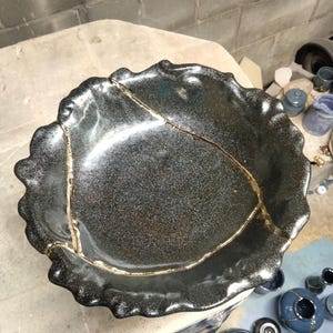 May include: A round ceramic bowl with a dark, metallic, speckled glaze and an irregular, wavy rim. The bowl features visible cracks repaired with a gold-colored material, creating a distinct pattern across the interior surface of the vessel.