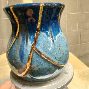 May include: A ceramic vase with a glossy finish featuring a gradient of deep navy blue to lighter sky blue tones. The surface is segmented by prominent, raised gold-colored lines that create a crackled, repaired appearance across the rounded body.