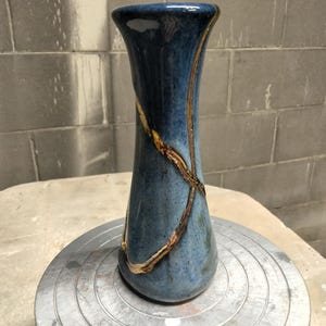 May include: A tall, slender ceramic vase with a glossy blue glaze featuring a mottled texture. The surface is decorated with prominent, raised gold-colored lines that wrap around the body in a crisscross pattern, highlighting the vessel's form.