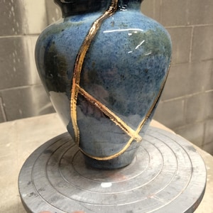 May include: A ceramic vase with a glossy blue and green mottled glaze, featuring prominent gold-colored repair lines that traverse the surface in a branching pattern, creating a distinct contrast against the dark, reflective ceramic body.