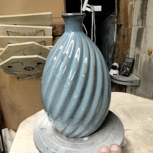 May include: A blue ceramic vase with a spiral design. The vase has a narrow neck and a rounded body. The color is a muted blue with darker accents in the grooves. The vase is sitting on a gray surface.