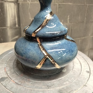 May include: A blue ceramic jar with a matching lid, featuring a glossy finish and irregular gold-colored lines that traverse the surface of both the body and the lid, creating a segmented appearance on the rounded, bulbous vessel.