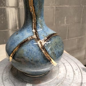 May include: A ceramic vase with a mottled blue and grey glaze featuring prominent gold-colored repair lines running vertically and horizontally across the body and neck, showcasing a kintsugi-inspired aesthetic with a smooth, reflective finish.