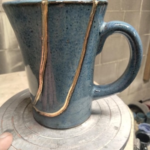 May include: A blue ceramic mug with a speckled finish and a curved handle. The exterior surface features a prominent, irregular vertical line filled with a metallic gold-colored material that contrasts against the textured blue glaze of the vessel.