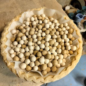 Peut inclure: Une croûte à tarte remplie de nombreuses petites billes rondes en céramique de différentes nuances de blanc et de marron. Les billes sont nichées sur une feuille de papier sulfurisé à l'intérieur de la croûte à tarte. La croûte a un bord décoratif festonné.