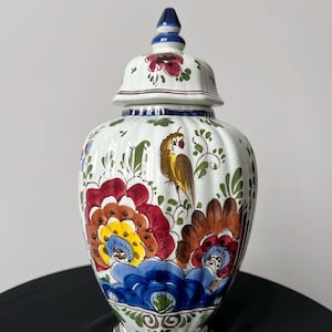 Könnte beinhalten: Eine weiße Keramikvase mit Deckel, verziert mit bunten Blumen- und Vogelmotiven. Die Vase hat eine gerippte Textur und einen dunkelbraunen Sockel. Der Deckel ist mit einer blauen Spitze versehen. Die Blumen sind in Rot-, Gelb- und Blautönen gehalten.