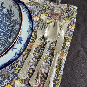 Può includere: Un'apparecchiatura con un piatto a motivi floreali blu e bianchi, posate e un tovagliolo decorativo. Le posate includono una forchetta, un cucchiaio e un coltello. Il tovagliolo ha un colorato motivo floreale e di uccelli. Sono visibili due calici di cristallo.