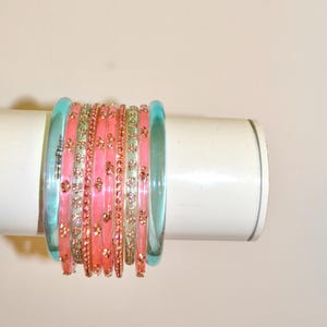 Può includere: Un set di dieci braccialetti impilati con due braccialetti azzurri traslucidi sui bordi esterni, alternati a braccialetti rosa e sottili fasce color oro decorate con piccoli accenti metallici ripetuti a forma di fiore.