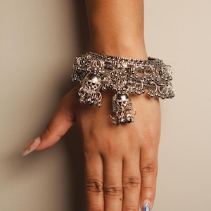 May include: A silver-toned metal bangle bracelet featuring an intricate textured band adorned with multiple small dangling bell-shaped charms and tiny spherical beads that hang from the main circular structure of the jewelry piece.
