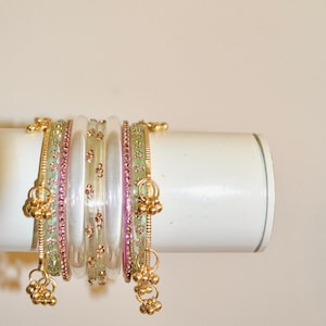 Op de afbeelding: Een set gestapelde armbanden met doorschijnende witte, lichtgroene en roze banden. De groene en roze banden zijn versierd met kleine bloemmotieven, terwijl de witte banden gouden accenten en hangende metalen bedels hebben aan de uiteinden.