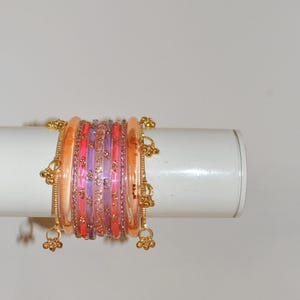 May include: A set of stacked bangles featuring translucent peach, pink, and purple bands. The set includes metallic gold-toned spacers with small floral patterns and dangling gold-toned charms shaped like tiny bells attached to the outer edges.