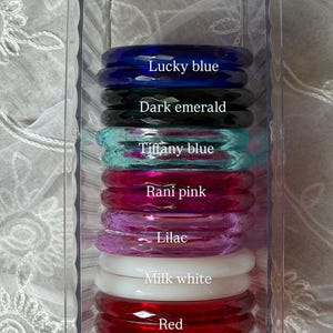 May include: A clear plastic rectangular storage container holding eight faceted bangles in various colors labeled Lucky blue, Dark emerald, Tiffany blue, Rani pink, Lilac, Milk white, Red, and Soft pink, arranged in a vertical stack.