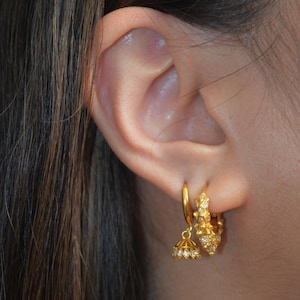 May include: Two gold hoop earrings worn in the earlobe. One hoop is plain and features a small, bell-shaped charm with tiny clear stones. The second hoop is thicker, textured with a spiked surface, and embedded with multiple small clear stones.