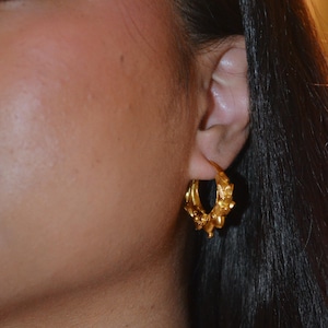 Pode incluir: Um close-up de um brinco de argola dourado com detalhes florais e de contas intrincados. O brinco é usado na orelha de uma pessoa com cabelo escuro. O brinco tem um acabamento polido.