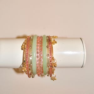 Könnte beinhalten: Ein Set aus gestapelten Armreifen mit abwechselnden Bändern in blassem Minzgrün, zartem Rosa und goldfarbenem Metall. Die rosa Bänder sind mit kleinen Blumenmustern verziert, während die goldenen Bänder zierliche Anhänger aufweisen.