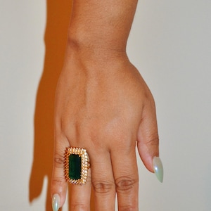 May include: A gold ring featuring a large, rectangular, emerald-green center stone surrounded by a halo of small, clear, sparkling stones. The ring is worn on the ring finger, showcasing a prominent, vibrant green gemstone set in a metallic band.