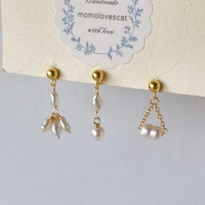 May include: Three pairs of handmade earrings with gold-colored posts. The earrings feature white pearls in various designs, including a floral cluster, dangling drops, and a chain-linked style. The earrings are displayed on a card with text that reads "Handmade momolovescat with love."