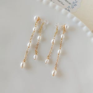 Può includere: Un paio di orecchini pendenti con piccole perle bianche infilate su delicate catene color oro. Gli orecchini sono a clip e il design include una piccola perla nella parte superiore e inferiore di ogni catena. Gli orecchini sono su un piatto bianco.