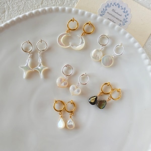 May include: A collection of earrings with various designs, including stars, moons, hearts, and teardrops. The earrings feature gold and silver hoops, and iridescent pearl-like pendants. The earrings are displayed on a white plate.