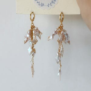 May include: A pair of gold-toned earrings featuring irregular, off-white baroque pearls. The earrings have a cascading design, with multiple pearls suspended from a gold wire. The top of the earrings have a gold hoop closure. The earrings are displayed against a light background.