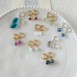 Puede incluir: Una colección de pendientes de piedras preciosas expuestos sobre un plato blanco. Los pendientes presentan varias piedras de colores, incluyendo azul, verde, morado y transparente, con aros dorados y plateados. Los pendientes están dispuestos en pares.
