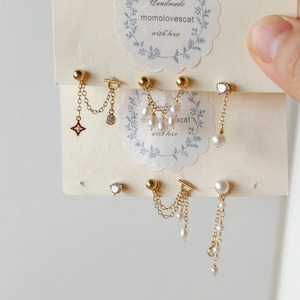 May include: A set of six gold-toned earrings displayed on a cream-coloured card with the text "Handmade momolovescat with love". The earrings feature delicate chains, small white pearls, and clear crystals in various dangling and stud designs.