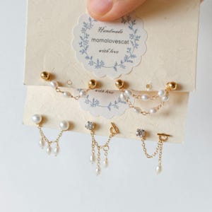 Puede incluir: Cinco pares de pendientes dorados presentados en tarjetas de color crema con el texto Handmade momolovescat with love. Los pendientes cuentan con pequeñas perlas blancas, delicadas cadenas doradas y piedras claras en varios diseños.