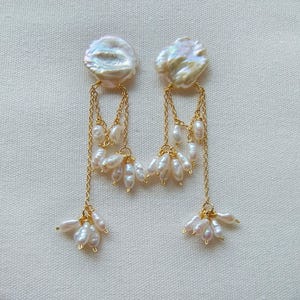 May include: A pair of dangle earrings with a luxurious design. Each earring has a large, iridescent, coin-shaped pearl at the top, connected by fine gold chains to clusters of smaller, teardrop-shaped pearls. The earrings have a floral design.