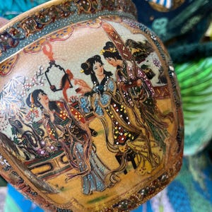 Puede incluir: Una maceta de cerámica con un borde dorado y detalles intrincados. La maceta presenta una escena de tres mujeres con ropa tradicional japonesa, pintadas en colores vibrantes con detalles dorados.