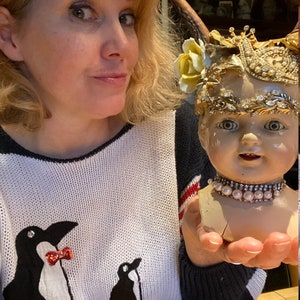 May include: A woman wearing a white and black sweater with penguin designs is holding a porcelain doll head with a gold crown and pearl necklace. The doll is wearing a gold crown with a yellow flower.