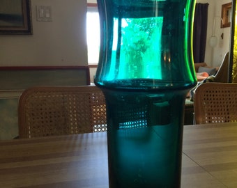 Könnte beinhalten: Eine hohe, blaugrüne Glasvase mit einer weiten Öffnung und einem abgerundeten Boden. Die Vase steht auf einem Holztisch.