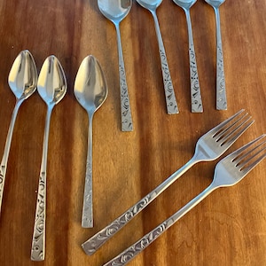 May include: A collection of stainless steel silverware, including spoons and forks, arranged on a wooden surface. The spoons have oval bowls and decorative etched handles. The forks have tines and matching etched handles. The silverware is likely for dining.