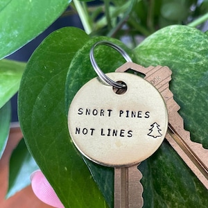 May include: A brass key ring with two keys and a round charm. The charm has the text "SNORT PINES NOT LINES" and a small tree graphic. The keys are attached to a silver ring, set against a backdrop of green leaves.