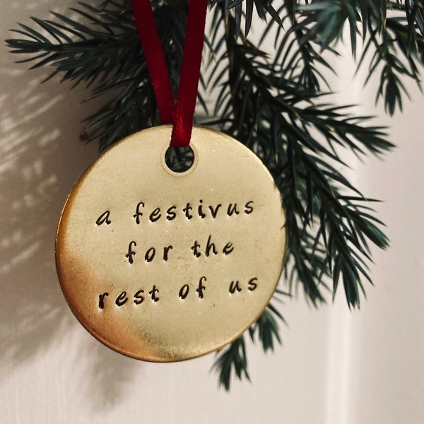 May include: A gold-coloured ornament with the text "a Festivus for the rest of us" hanging from a red ribbon on a green pine branch.