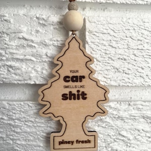 May include: Wooden car air freshener in the shape of a pine tree. The text on the freshener reads "YOUR car SMELLS LIKE shit" and "piney fresh". The freshener is attached to a brown string with a wooden bead.