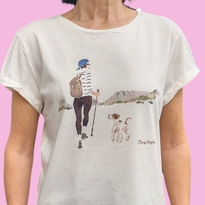 Puede incluir: Camiseta de color crema con una ilustración en acuarela de una persona caminando con un perro y bastones de senderismo. La persona lleva un gorro azul, una camisa a rayas y una mochila. El perro es blanco y marrón. La firma del artista es visible.