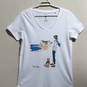 May include: White v-neck t-shirt with a watercolor illustration of a person walking a dog on a beach with a cliff in the background. The text "Elena Copley" is printed on the shirt.