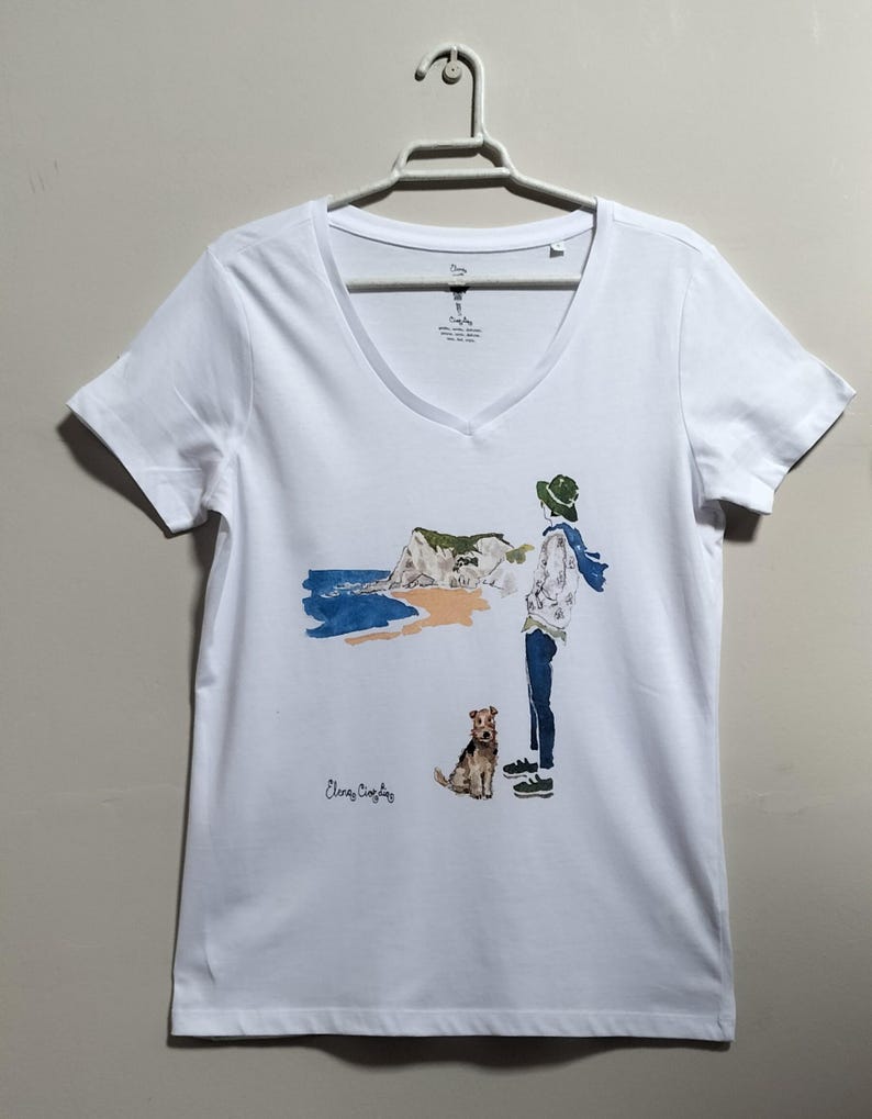 May include: White v-neck t-shirt with a watercolor illustration of a person walking a dog on a beach with a cliff in the background. The text "Elena Copley" is printed on the shirt.