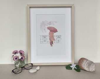 May include: A framed watercolor painting of two people walking under a red umbrella in the rain. The painting is in a light wood frame and is set on a table with a vase of flowers, a pair of glasses, and a rock.