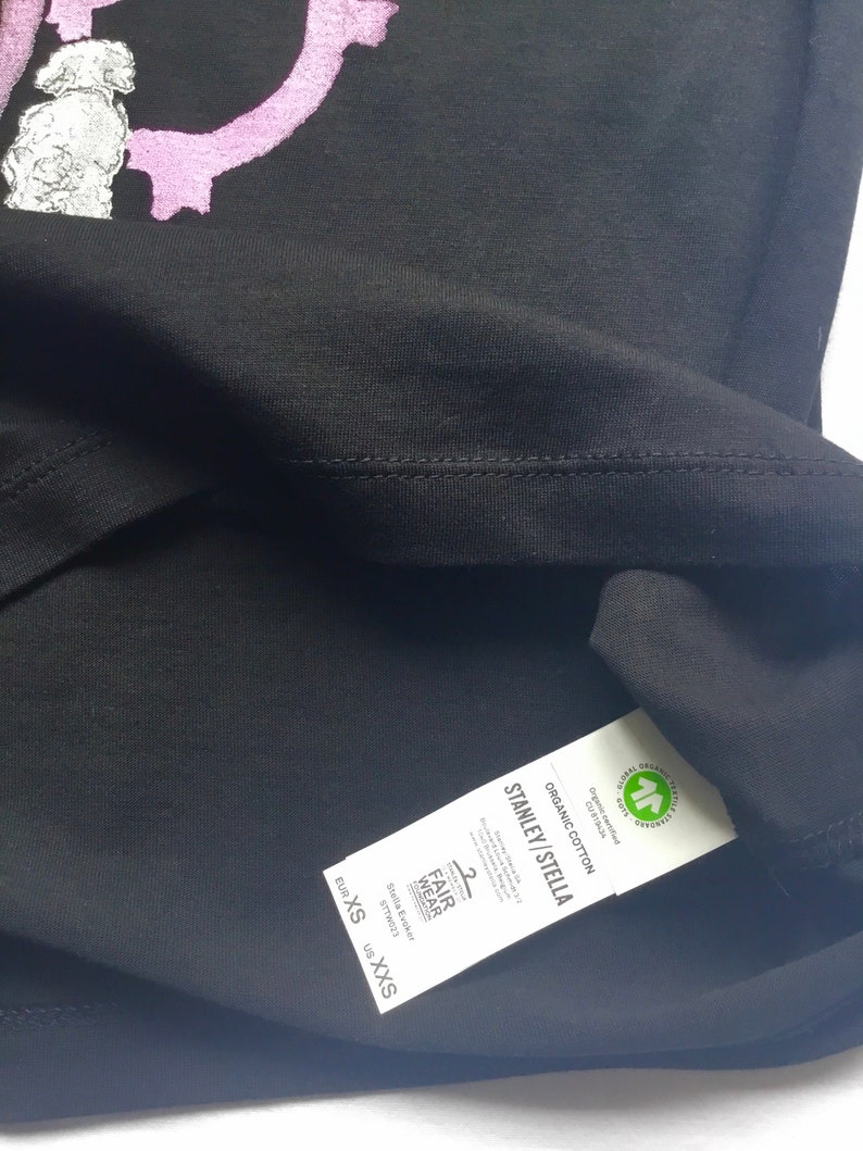 May include: Black t-shirt with a pink graphic print. The tag reads "Stanley/Stella" and "Organic Cotton" with a Global Organic Textile Standard (GOTS) certification logo. The size tag reads "EUR XS, US XXS".