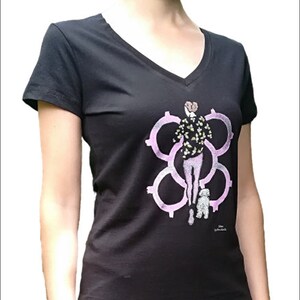 May include: Black v-neck t-shirt with a pink and green graphic print of a woman walking a small white dog. The graphic features a stylized design of interlocking circles.